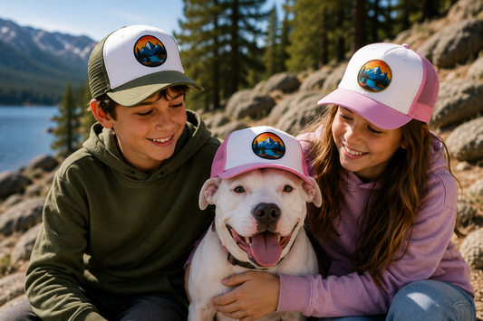 A boy and a girl outdoors on rocky terrain by a lake, each wearing a white trucker hat with a pink brim and a circular mountain logo. A white dog is sitting in the center wearing a matching pink-trimmed hat.